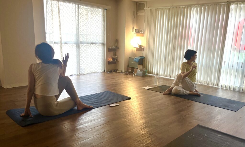 女性2名が渋谷にあるNew York Style YOGAのスタジオで、Sivananda Yoga のクラス中、背骨をねじる座位のヨガのポーズをとっている